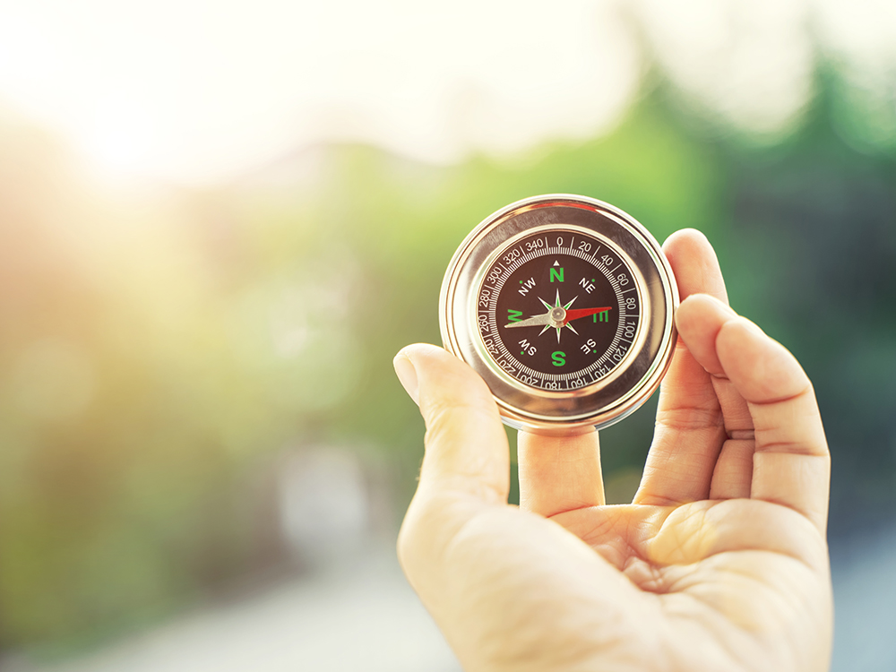 Closeup of hand holding compass with blurred green nature and sunlight with flare background with free copy space for text. Life, travel and guide for success concept.